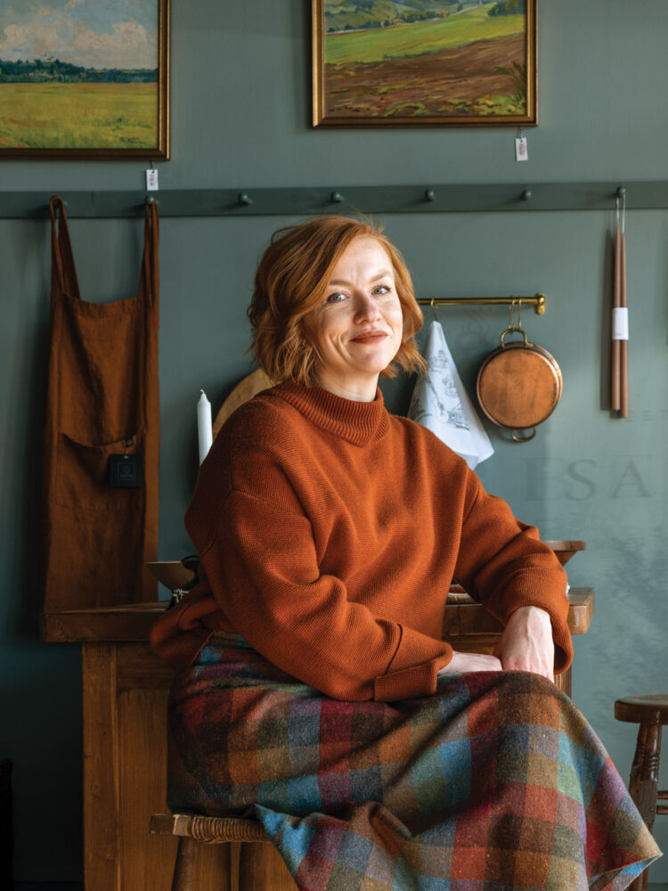 A red-haired woman sits in her dark teal storefront in an orange sweater and flannel skirt.