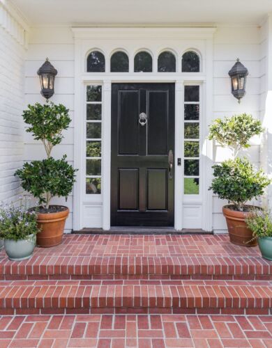 Black, four-panel front door with sidelights and arched transom lights, flanked by pair of potted evergreens on brick landing. 