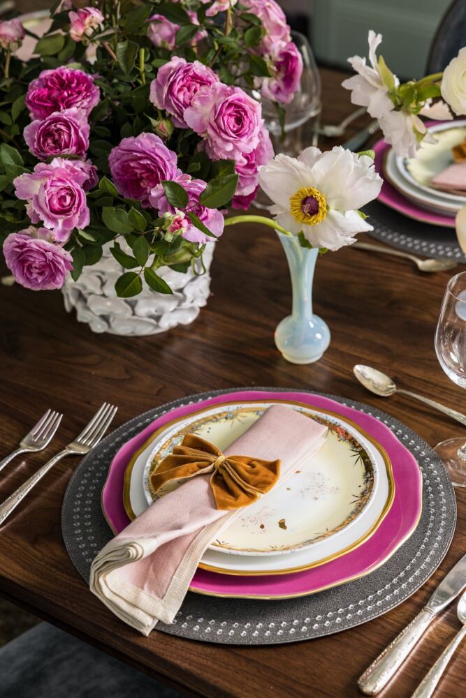White ceramic vase of pink roses on dining table with pink, gold, and gray place setting with gold bow napkin ring on rose-colored napkin.
