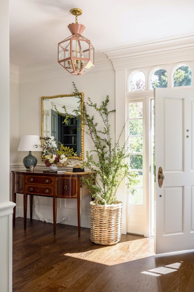 Sunny foyer with open door and antique sideboard topped with gold Carver's Guild mirror, handmade ceramic lamp, and vase of flowers and foliage branches. A woven basket with tall green branches stands beside the table.