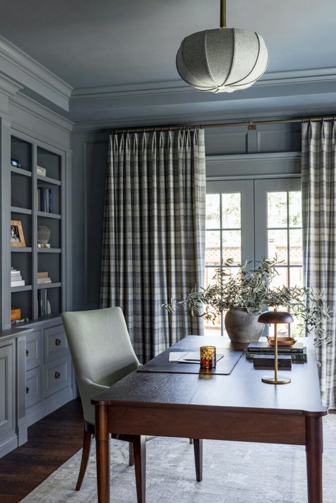 Home office with bookcases, walls and ceiling painted in a shade of navy blue. Desk topped with reading lamp and rustic ceramic jar of olive branches.