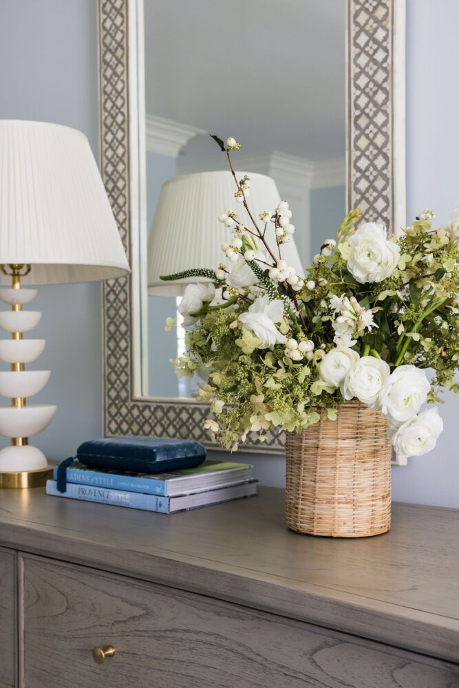 Bedroom dresser topped with mod lamp, stack of books, and rattan-covered vase of white ranunculus, green-tinged oakleaf hydrangea blossoms, white berries, and white tuberose flowers.