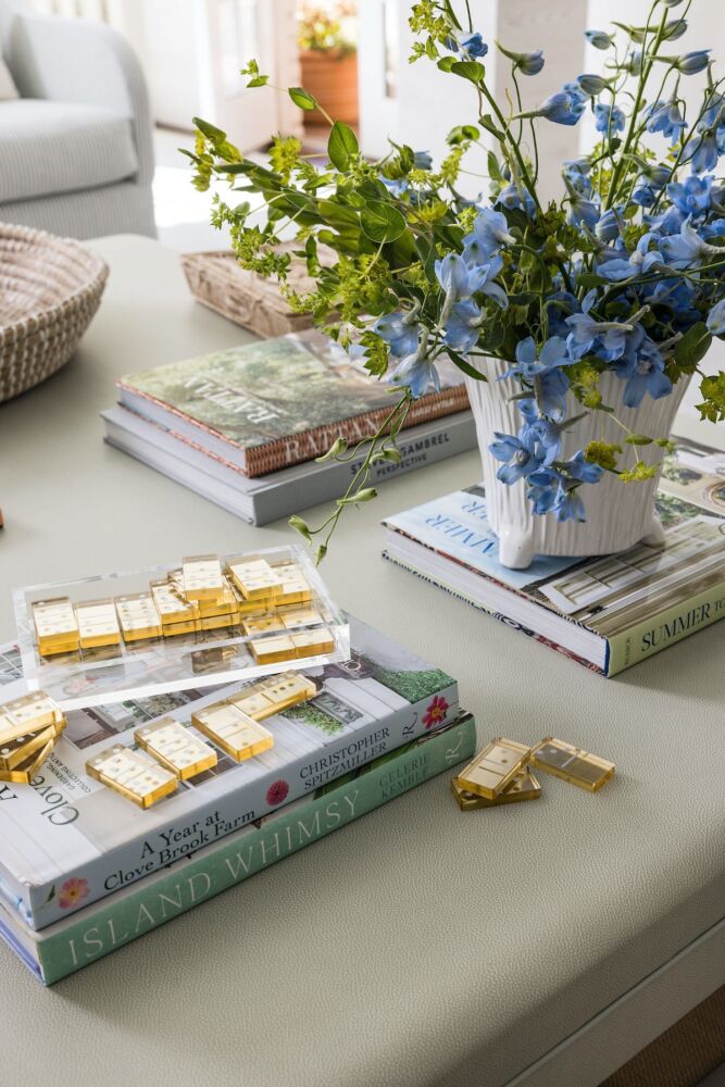 Stacks of books, a white ceramic vase filled with blue delphinium and belles of Ireland, along with amber-toned dominoes top the living room coffee table.