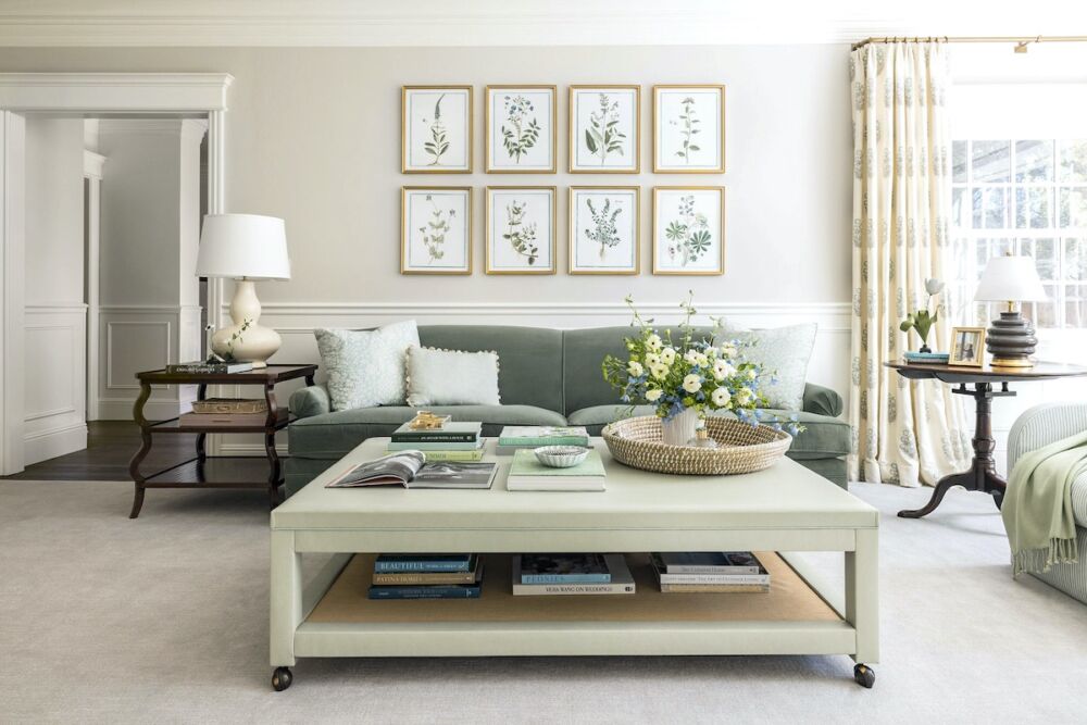 Large, leather-covered coffee table on casters in living room with sage grey sofa and antique side tables. Botanical prints on wall above sofa.