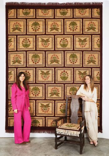 A floral rug hangs on the wall behind two women.