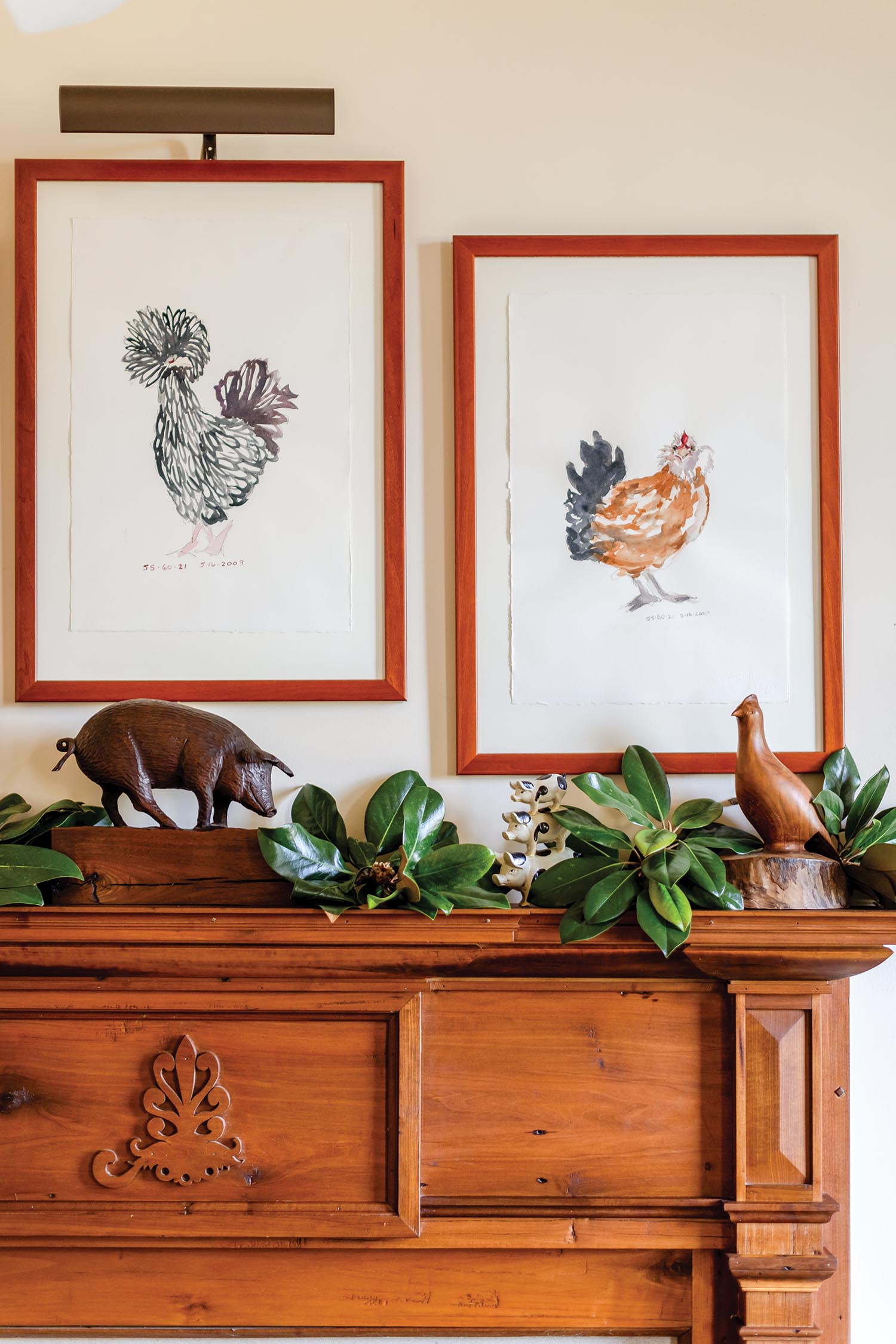 Two red-framed watercolored chickens hang above a wooden mantel holding magnolia leaves.