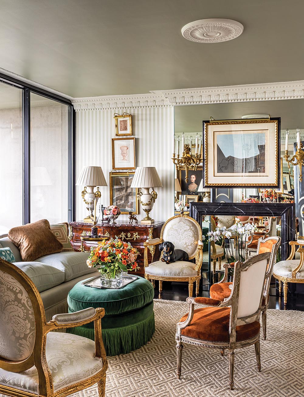 A living room has grey striped wallpaper, antiques, floral arrangements, and a dog.