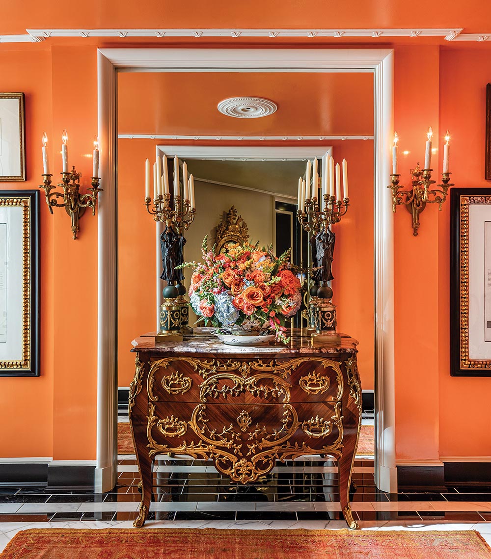 A floral arrangement sits on top of a French antique in an orange hallway.