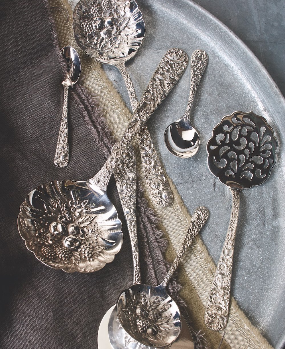 Repousse silver serving spoons: berry, bonbon, and nut spoons—in all three sizes—include a charming pattern of fruit and flora in the bowl. A small one sits on a serving piece with a completely flat bowl used for hotcakes. The tomato spoon is also flat with a pierced bowl, and the smaller spoons are for demitasse and for baby.
