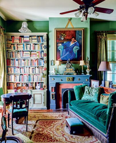 A green room with a portrait hanging over a fireplace, a stuffed bookshelf, and a velvet couch.