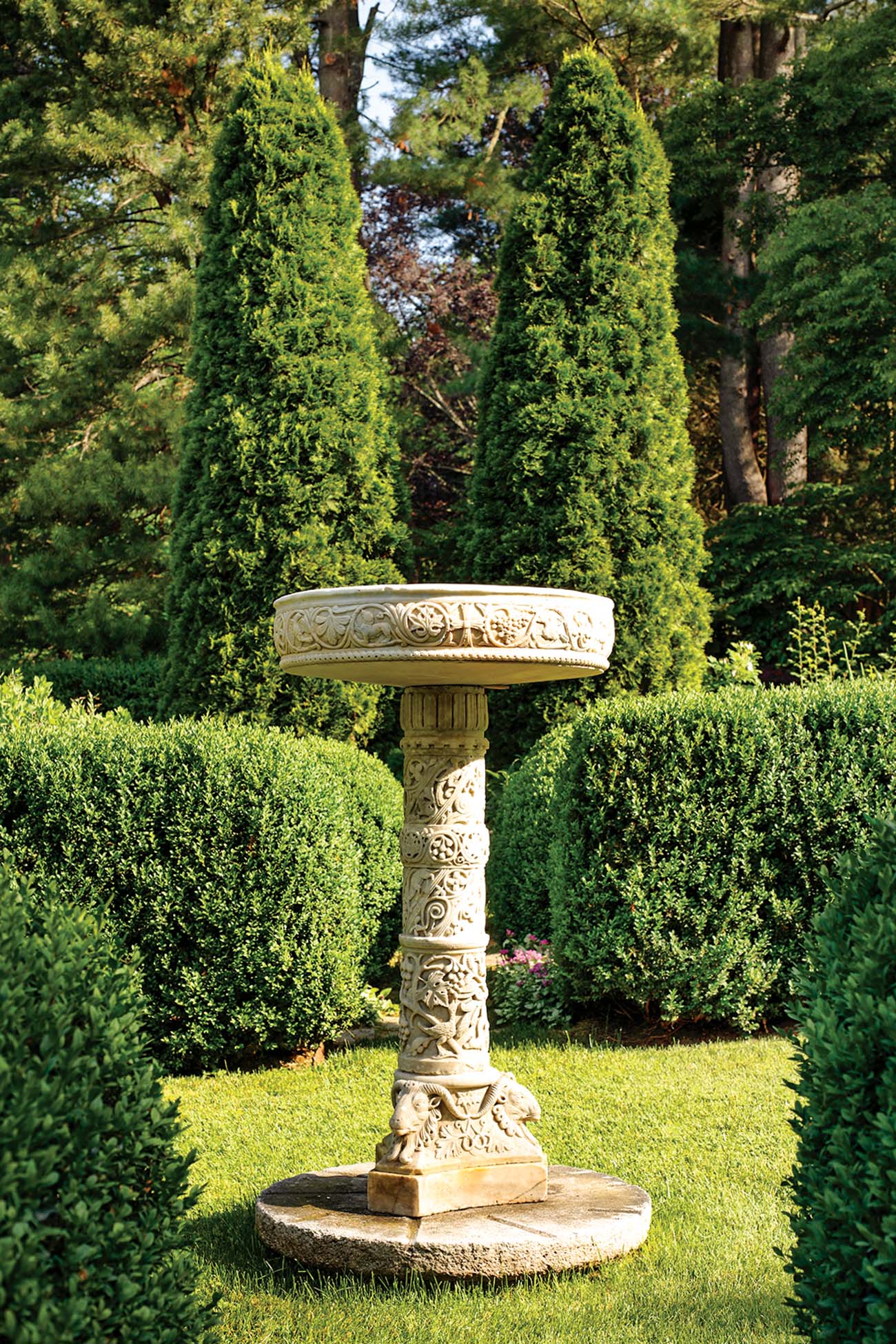 A stone basin sits in a garden next to manicured hedges.