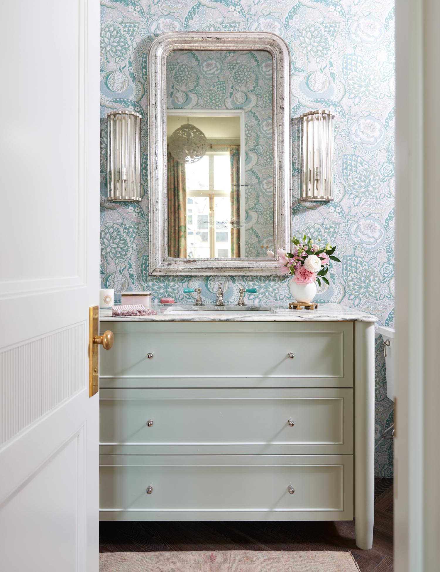 A bathroom has light blue wallpaper and a pastel teal dresser sink.