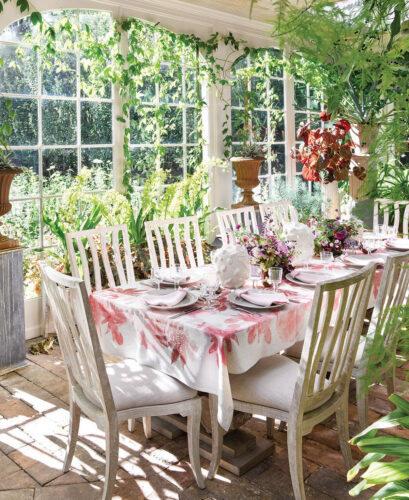 A long table with a pink and white floral tablecloth sits in a sunroom decorated with hanging greenery.