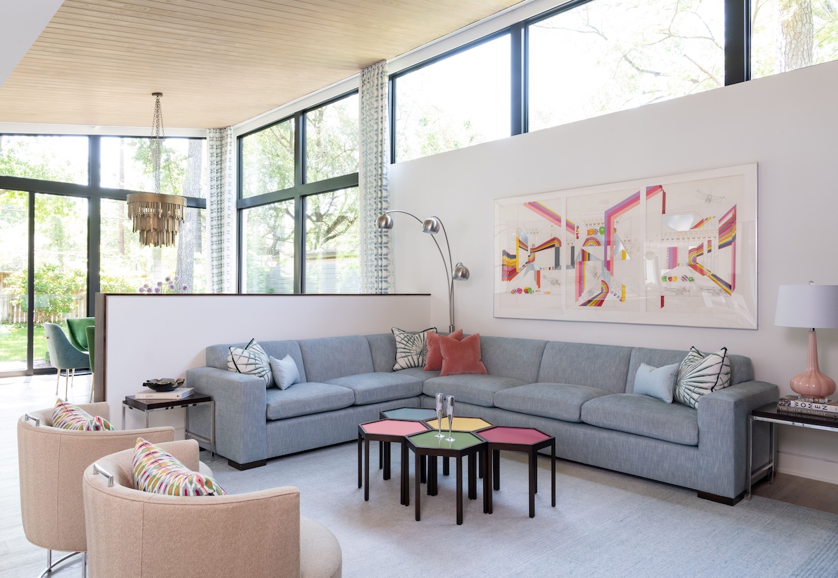A contemporary sectional sofa serves as the focal point for a bright and airy living room. Custom-painted hexagonal bunching tables, a bright piece of art, and accent pillows add colorful details to a space grounded by monochromatic blues.