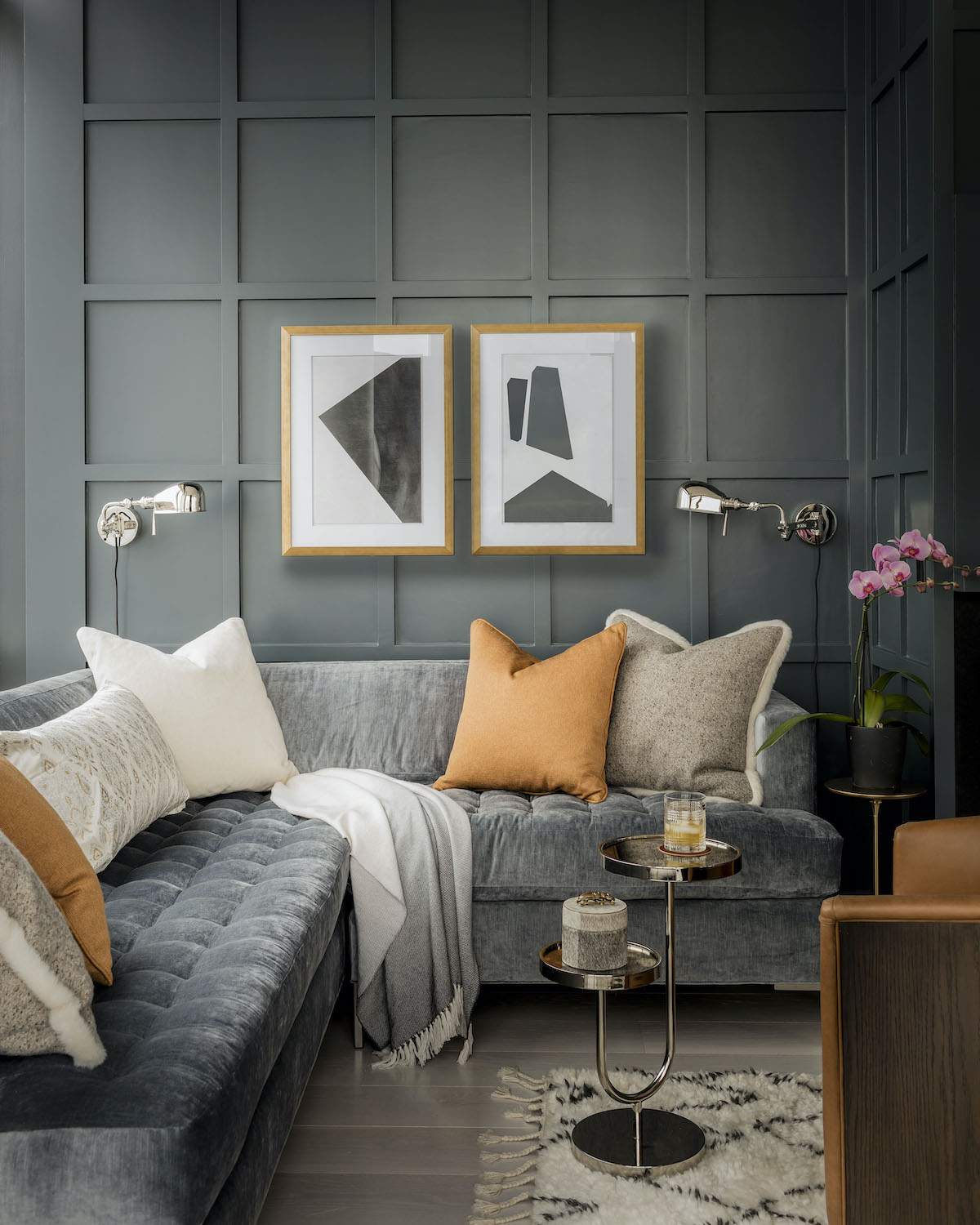 Grey sectional sofa with biscuit tufting on the seat to mirror the rectilinear paneling on the walls. Pair of silver sconces flank pair of lithographs on wall.