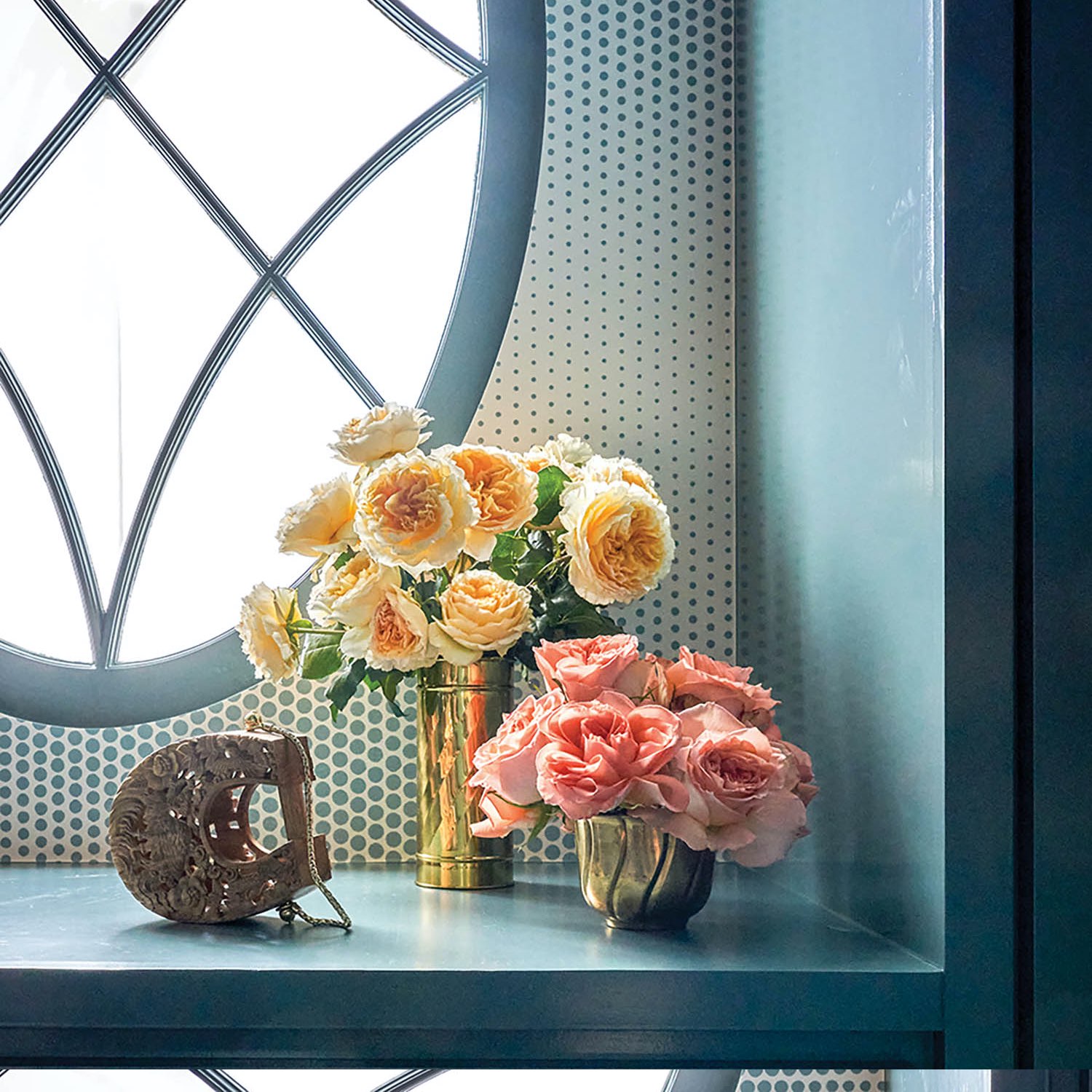 Yellow and pink roses in brass vases in front of an oval window.